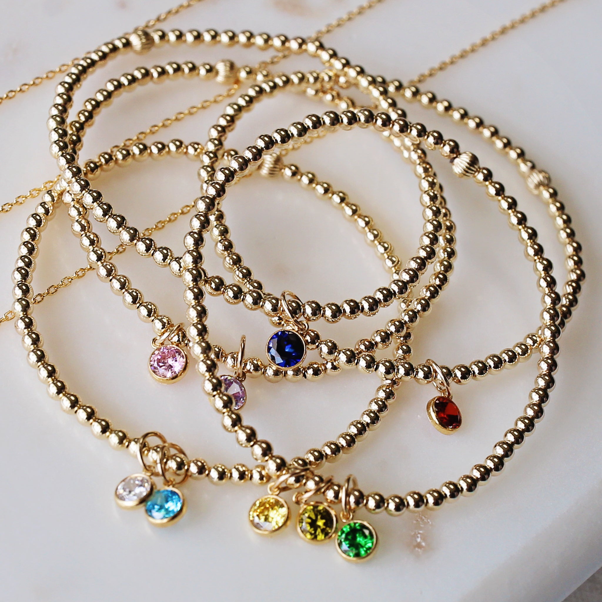 A gold-filled bracelet with beaded design featuring a charm with a birthstone, and additional charms in blue and red colors.