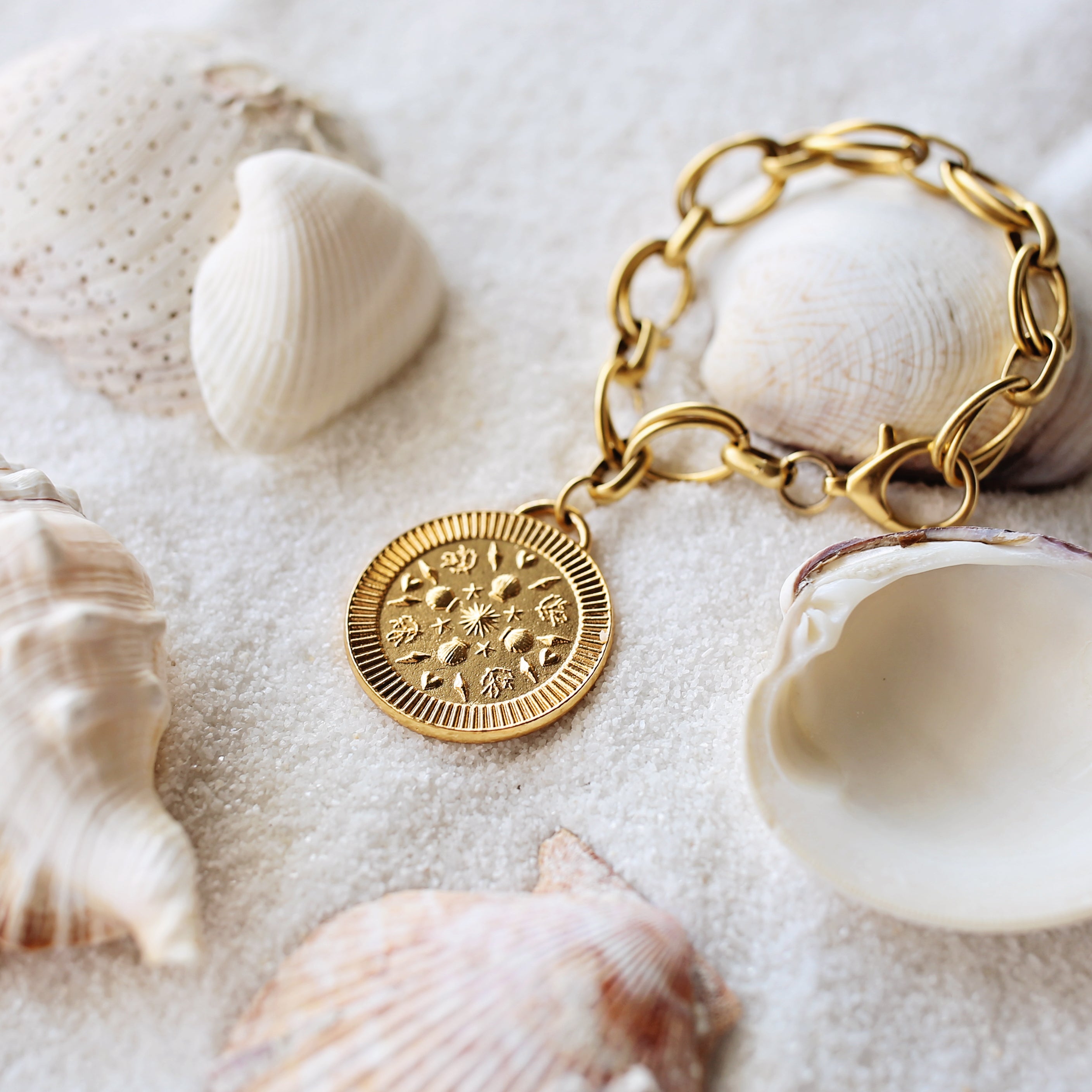 Gold bracelet with a decorative charm on a sandy surface with seashells.