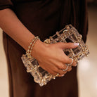 Hand holding a clear clutch with gold accents, wearing a gold bracelet and ring.