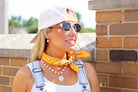 Woman wearing a yellow bandana, sunglasses, and overalls against a brick wall.